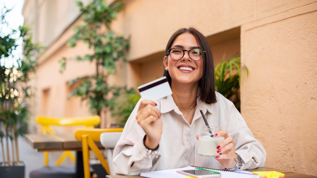 Smiling woman holding credit card and drink at outdoor cafe