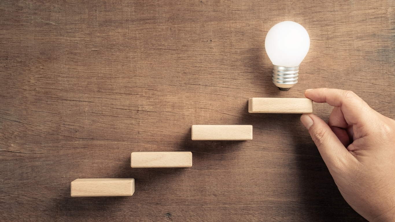 Hand building staircase toward glowing light bulb, symbolizing progress and ideas