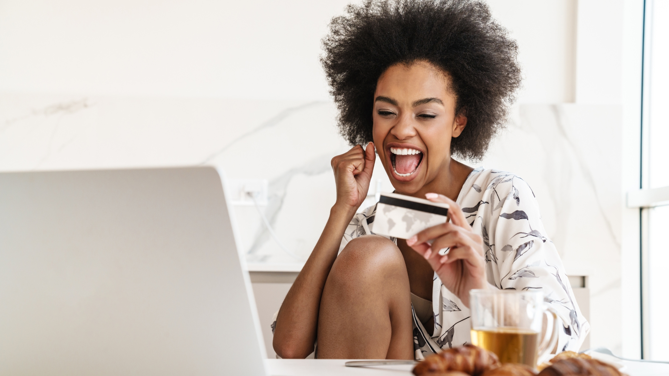 Excited woman shopping online with credit card and laptop, enjoying coffee at home.