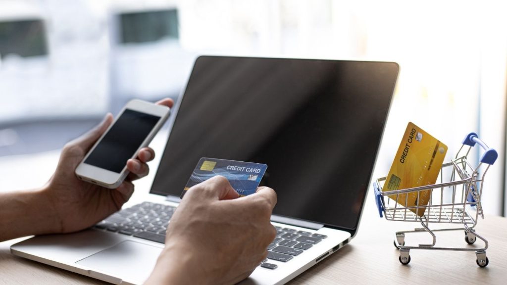 Person holding a smartphone and credit card in front of a laptop with mini shopping cart on desk, Rebuild Credit with Low Score