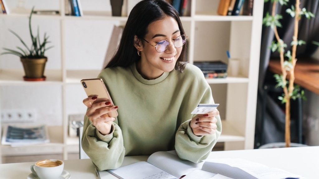 Young woman using smartphone and holding a credit card while studying at home