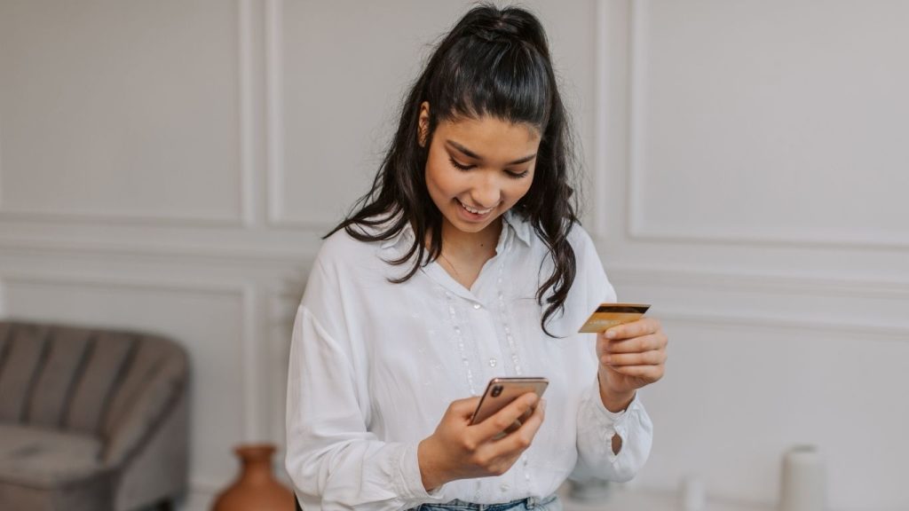 Young woman smiling while using smartphone and holding a credit card indoors