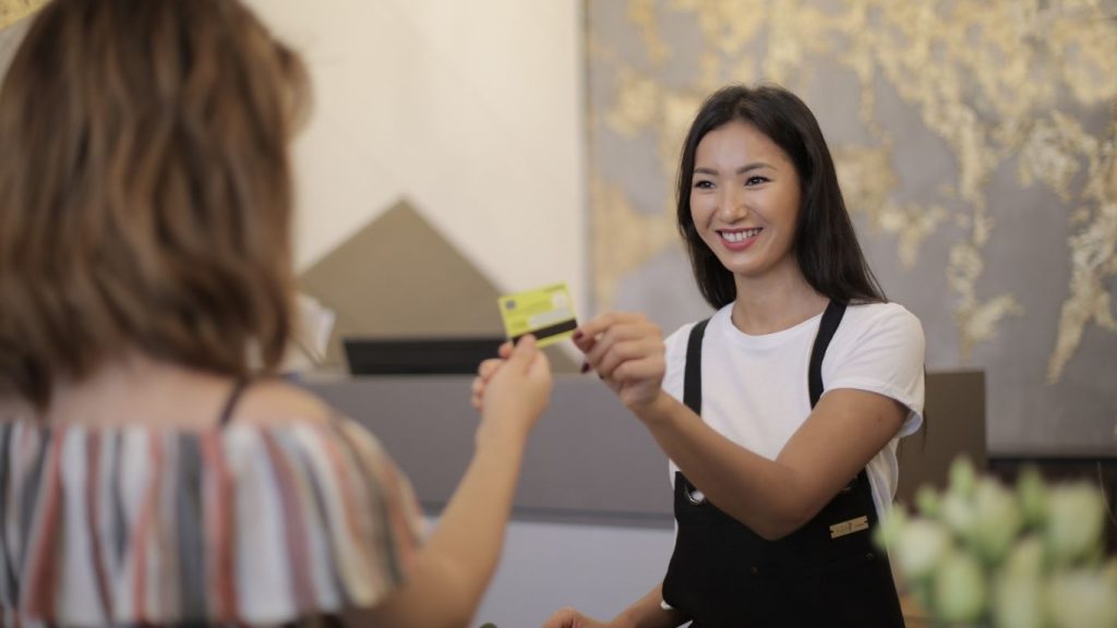 A smiling receptionist hands back a credit card to a customer at the front desk. Fix Credit to Improve Finances.