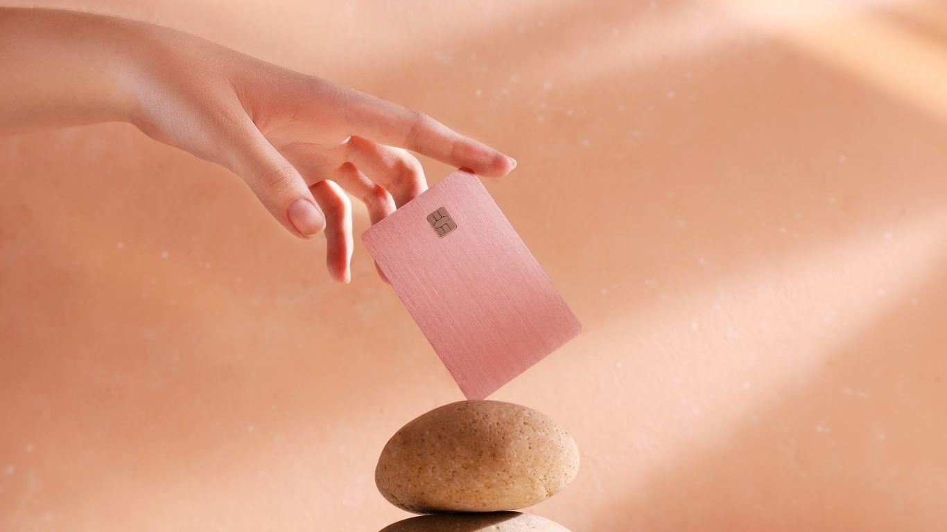 Hand placing a sleek pink credit card on a smooth stone against a soft peach background.