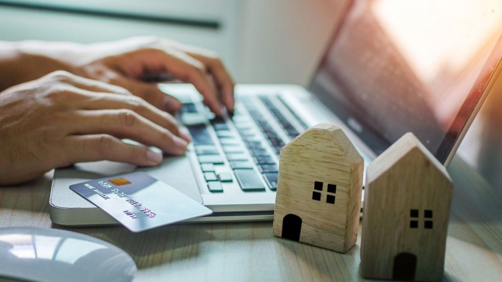 Hands typing on a laptop with a credit card and miniature wooden houses on the desk.Credit Repair for Homeownership.