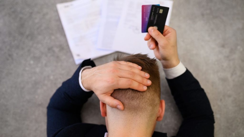 A stressed man holding credit cards with overdue bills on the table, symbolising credit card debt and financial pressure.