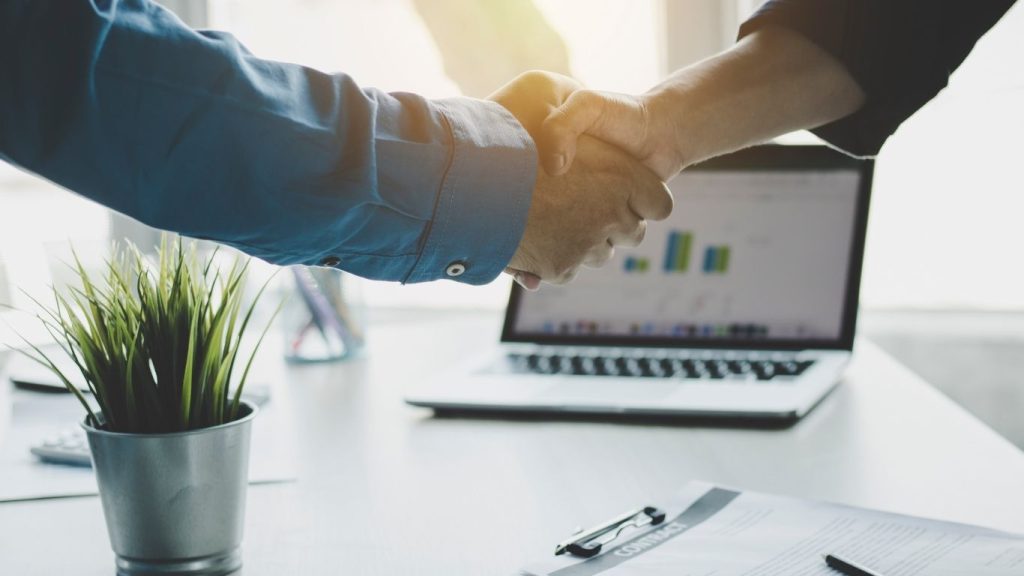 Two professionals shaking hands in an office with laptop and documents in background, symbolizing agreement or partnership.