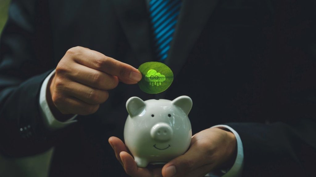 Businessman placing a green coin into a white piggy bank, symbolizing saving money and financial planning.