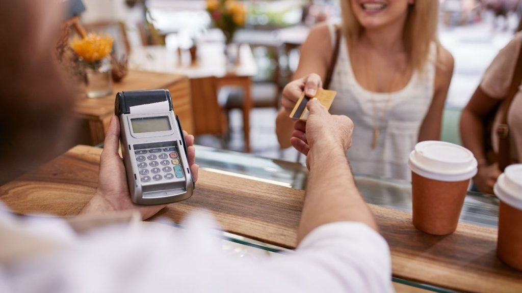 A smiling customer hands a credit card to a barista at a coffee shop counter, suggesting a smooth and confident payment experience.