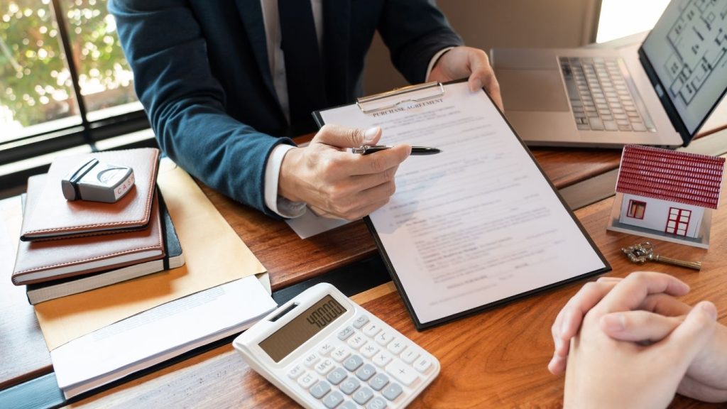 A financial advisor reviewing a document with a client, with calculator, house model, and documents on the desk—symbolizing loan or mortgage consultation.