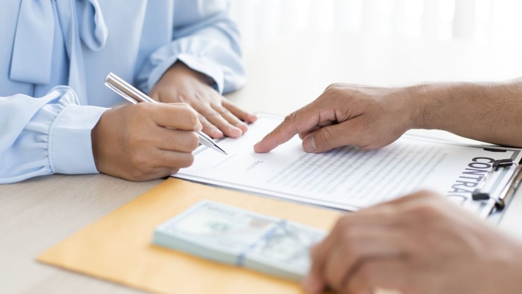 A person signing a financial contract while another hand points at the document, with a stack of dollar bills and an envelope on the table. Credit stress