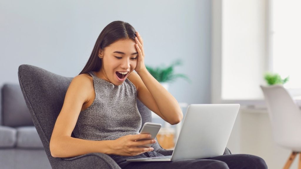 A young woman looks excited while checking her smartphone with a laptop open in front of her.