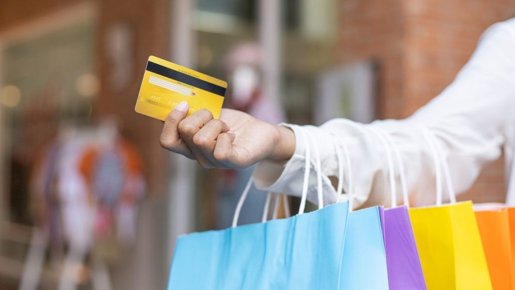 A person holding a yellow credit card and colorful shopping bags, standing in a shopping area.